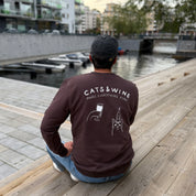 Person wearing a brown long-sleeve shirt with 'Cats & Wine Make Everything Fine' text and graphics, sitting on a wooden dock by a waterfront.